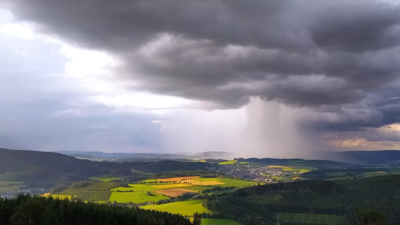 Dunkle Gewitterwolken mit starkem Regenschauer über einer ländlichen Landschaft mit Feldern und kleinem Ort.
