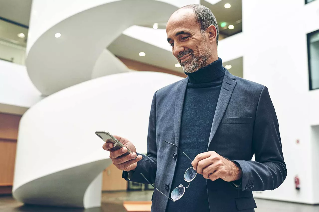 lächelnde-person-im-sakko-schaut-auf-smartphone-und-hat-brille-in-der-hand