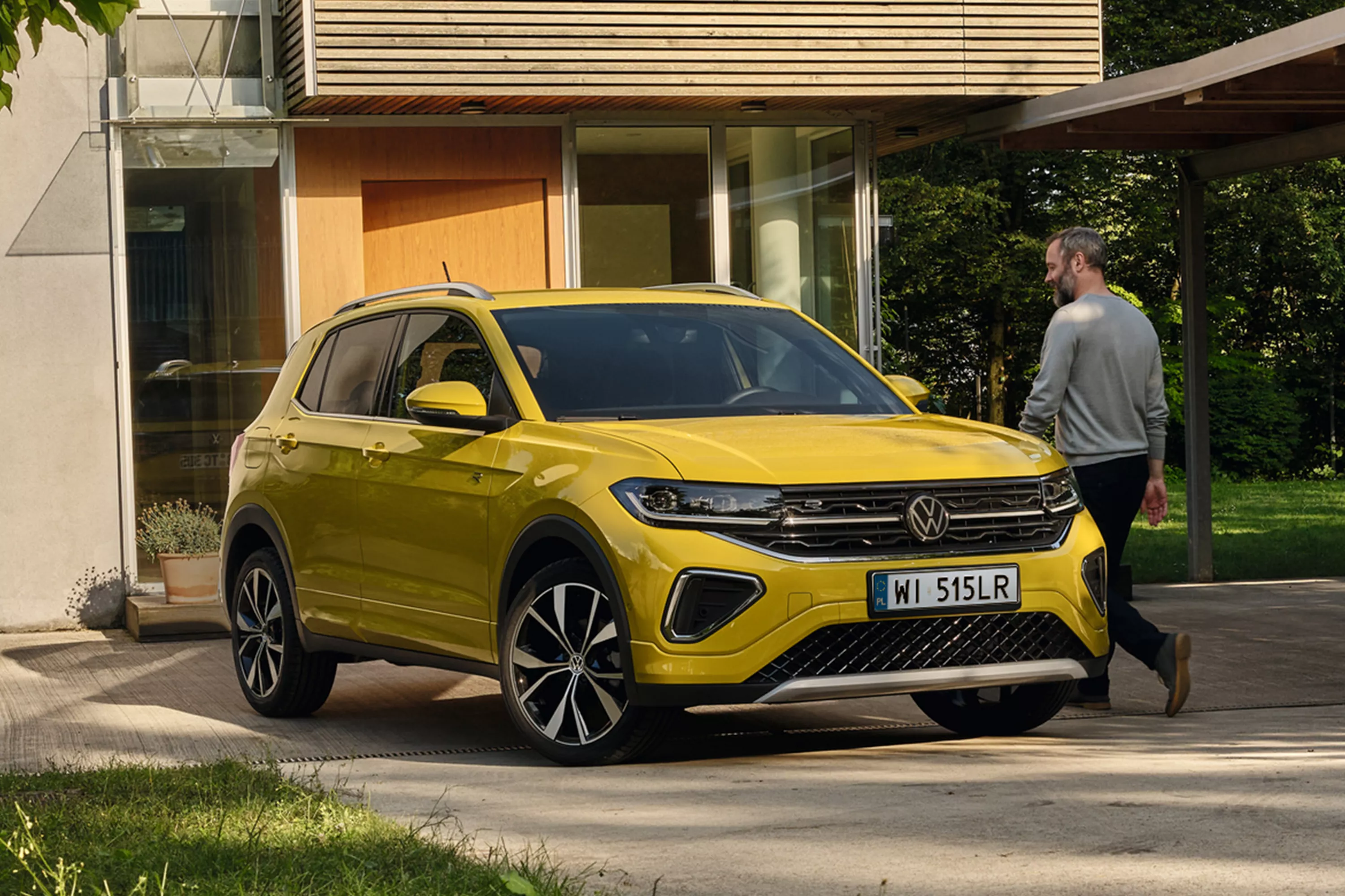 Żółty Volkswagen T-Cross przed nowoczesnym przeszklonym domem, w otoczeniu zieleni, obok auta idzie mężczyzna.   