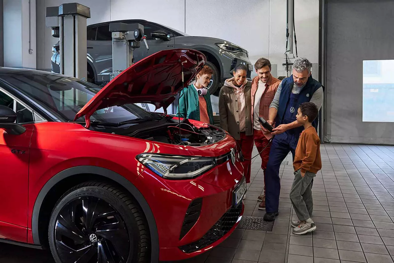 Werkstattmitarbeiter erklärt einer Familie den geöffneten Motorraum eines roten VW-Elektroautos in der Werkstatt
