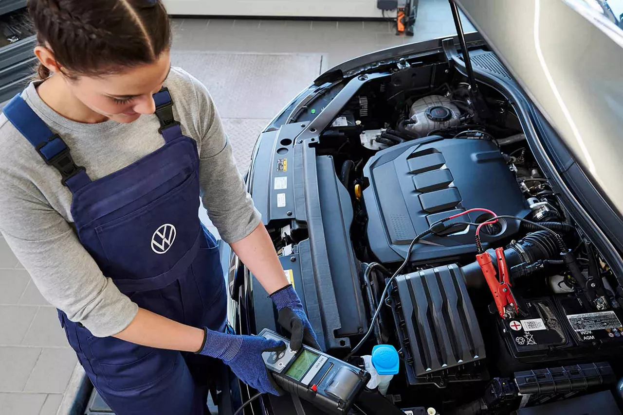 Mechanikerin im VW-Werkstattanzug prüft die Batterie eines Autos mit einem Diagnosegerät bei geöffneter Motorhaube
