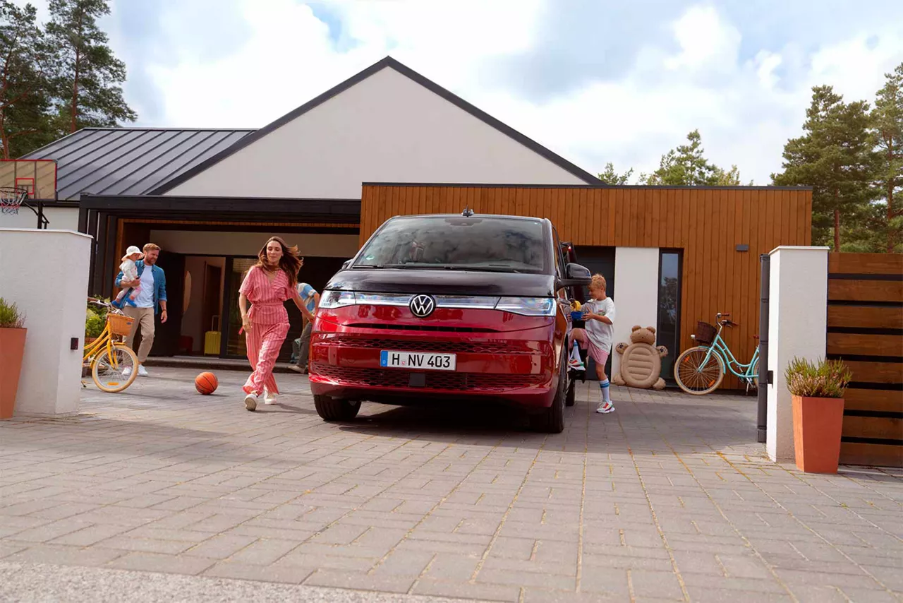 Roter Volkswagen Multivan steht in einer Einfahrt vor einem modernen Haus, mehrere Personen und Fahrräder im Hintergrund.