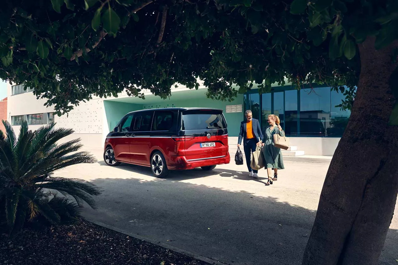 Roter VW Multivan steht vor modernem Bürogebäude, zwei Personen mit Taschen gehen Richtung Fahrgzeug.