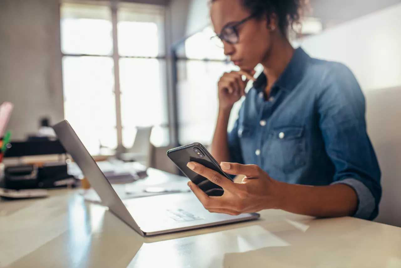 Eine Geschäftsfrau sitzt an ihrem Laptop und hat ihr Handy in ihrer Hand und schaut auf das Preis- und Leistungsverzeichnis