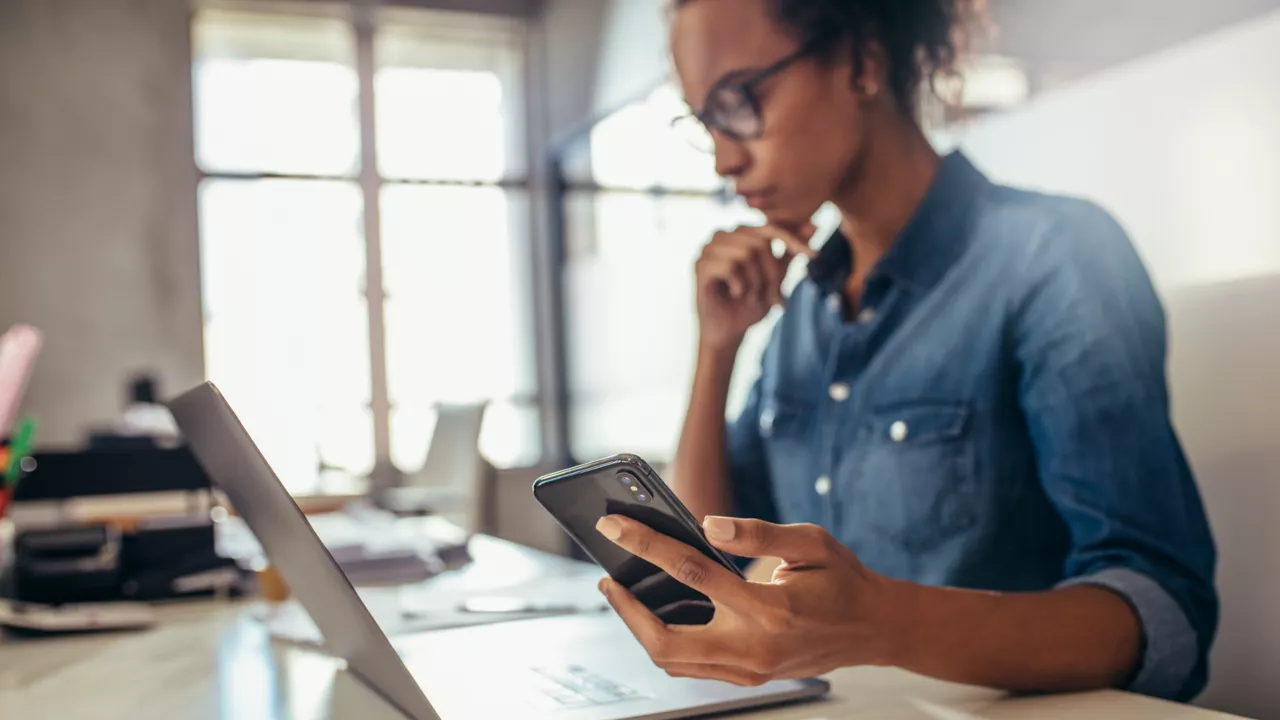 Eine Geschäftsfrau sitzt an ihrem Laptop und hat ihr Handy in ihrer Hand und schaut auf das Preis- und Leistungsverzeichnis