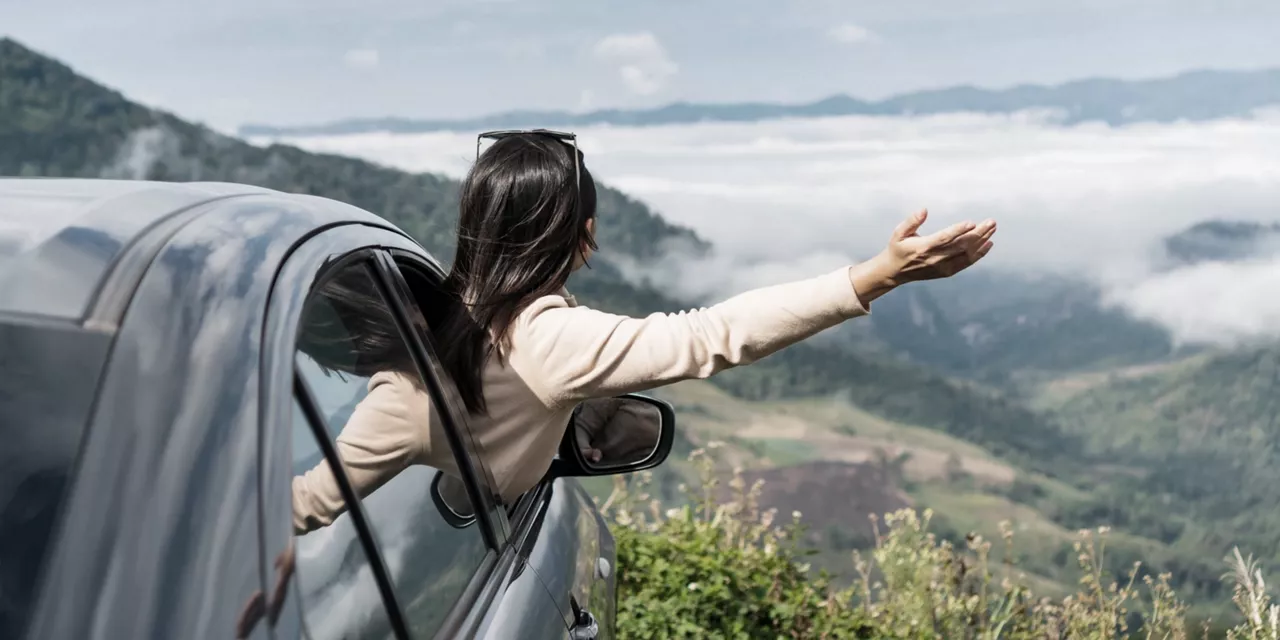 Mujer de espaldas sacando el brazo por la ventanilla de un coche estacionado en la naturaleza.