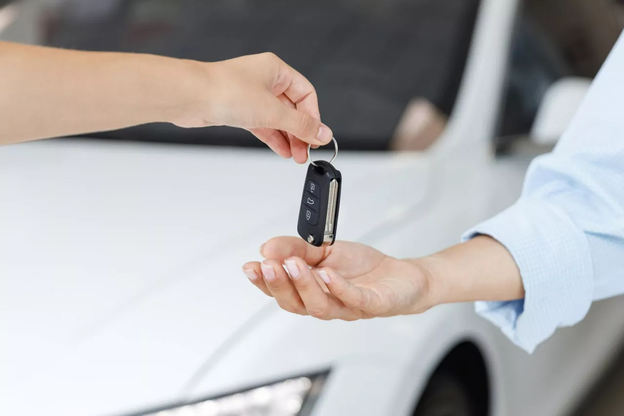 Zwei Arme übergeben sich einen Autoschlüssel. Im Hintergrund steht ein weißes Auto.