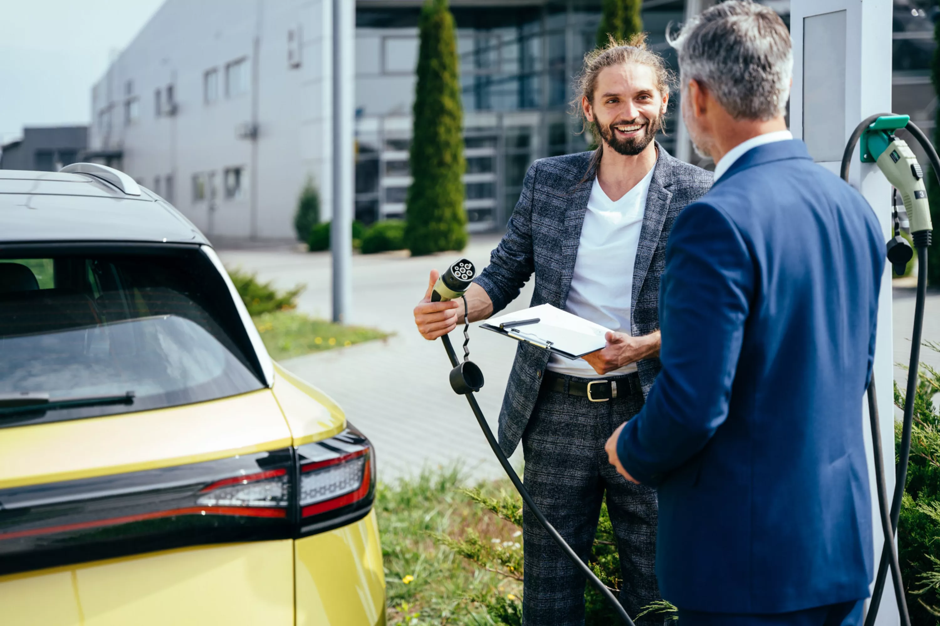 Zwei Geschäftsmänner neben E-Auto unterhalten sich über die Fuhrparkmanagement-Aufgaben