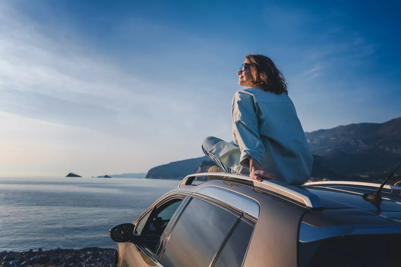 Person sitzt auf Ihrem Autodach. Es steht an einem Steinstrand mit dem Blick richtung Meer.