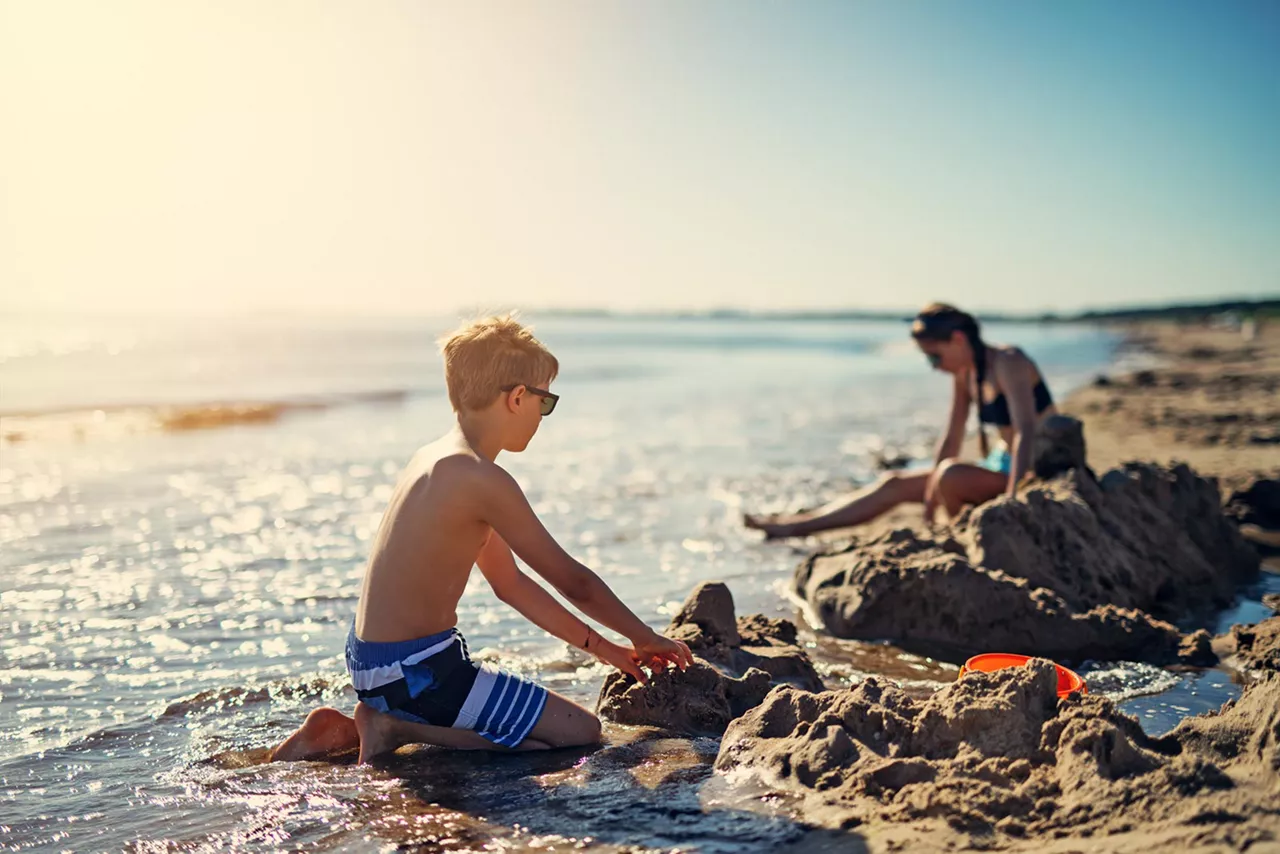 Kind sitzt am Wasser und baut Sandburgen bei Sonnenschein; Frau im Hintergrund