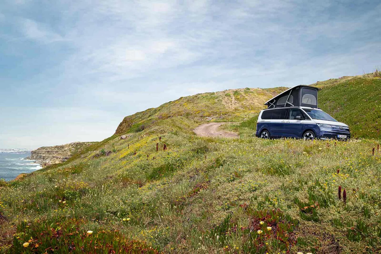 Blauer VW California steht mit offenem Aufstelldach auf einer Düne mit Meer im Hintergrund