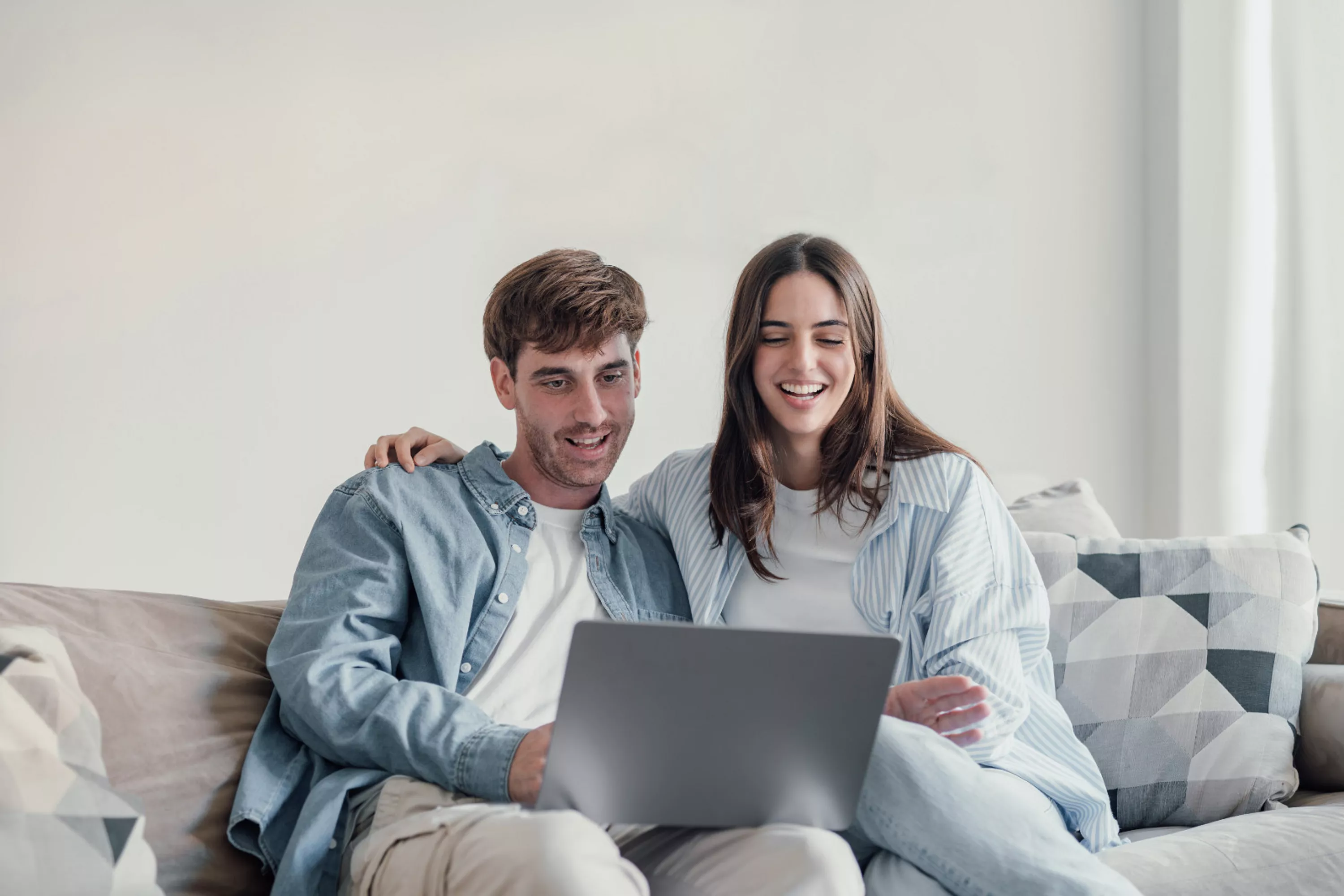 Ein junger Mann und eine junge Frau sitzen auf einem Sofa, das vor einer hellen Wand steht. Der Mann hat einen Laptop auf seinem Schoß, auf dessen Bildschirm beide schauen.