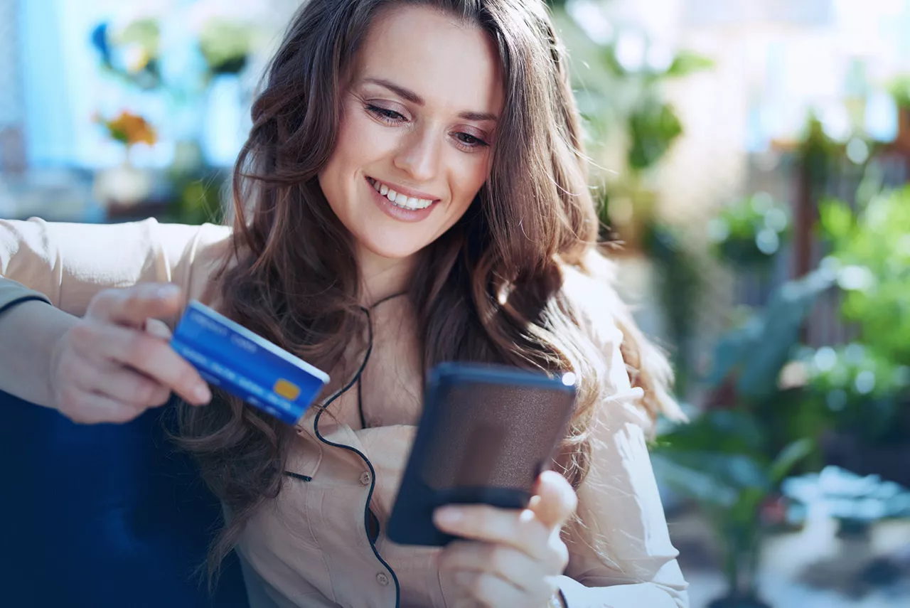 Uśmiechnięta kobieta patrzy w ekran telefonu, trzymając kartę bankową w drugiej ręce.