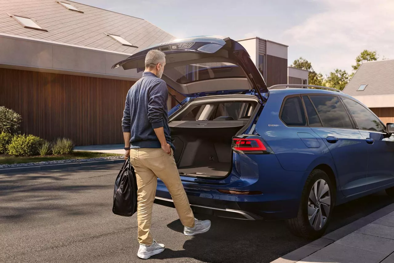 Person steht mit Aktentasche vor einem offenen Kofferraum eines blauen VW Golf Variant