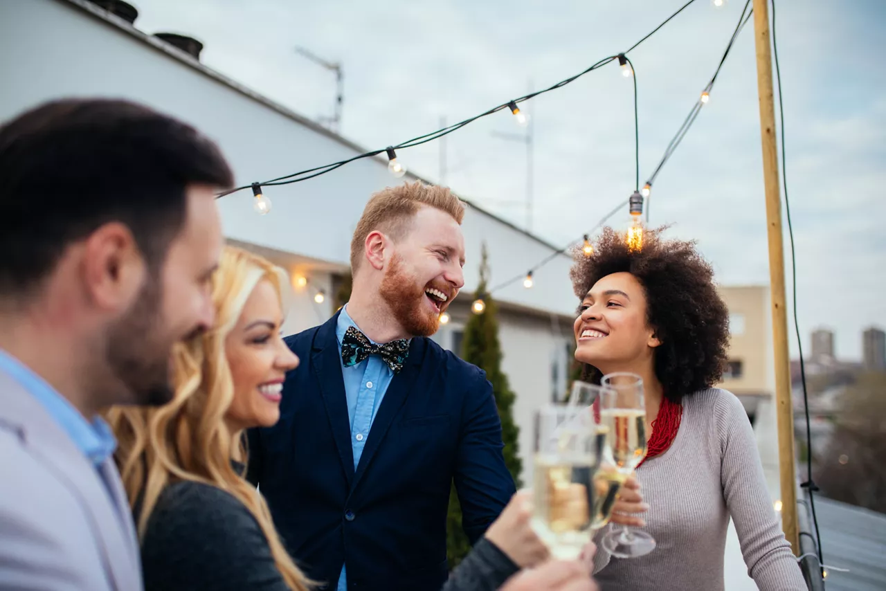 Seitliche Ansicht von mehreren Personen, die lachend mit einem Sekt in der Hand auf einem Dach stehen.
