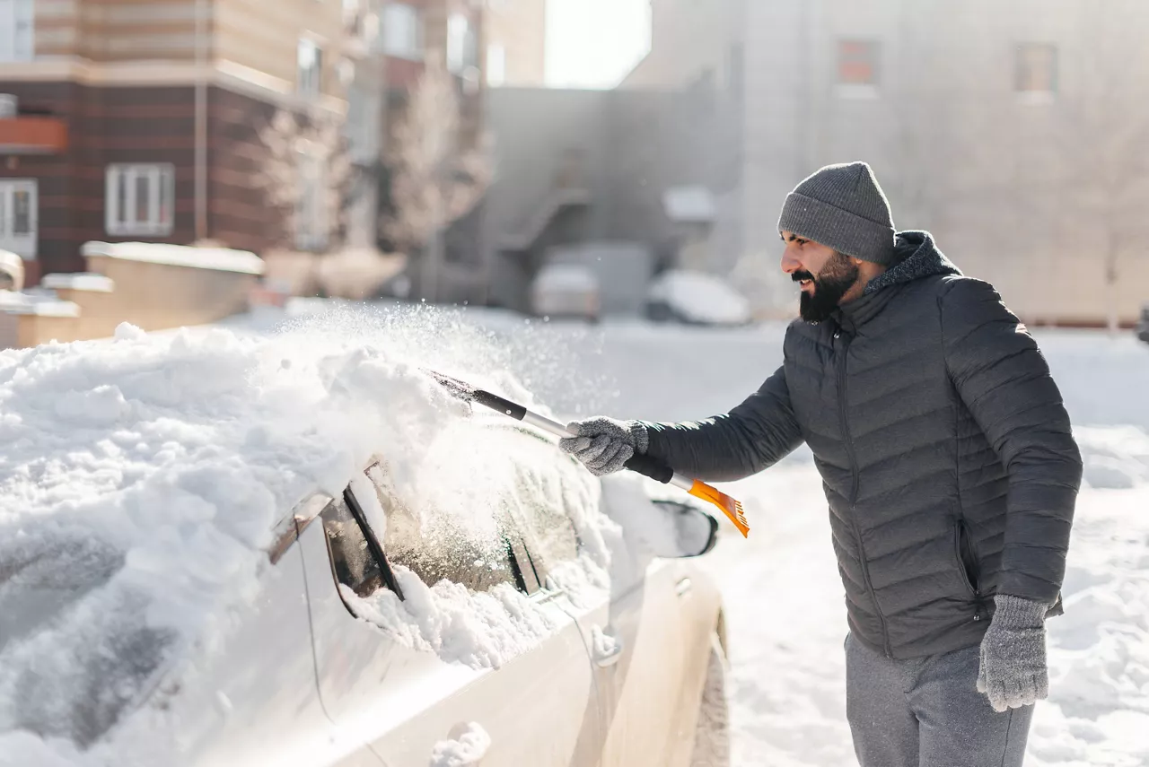 Ein Mann in Winterkleidung, mit Mütze und Handschuhen bekleidet, entfernt mit einem Werkzeug Schnee vom Dach eines Autos. Im Hintergrund sind Gebäude und Schnee zu sehen.