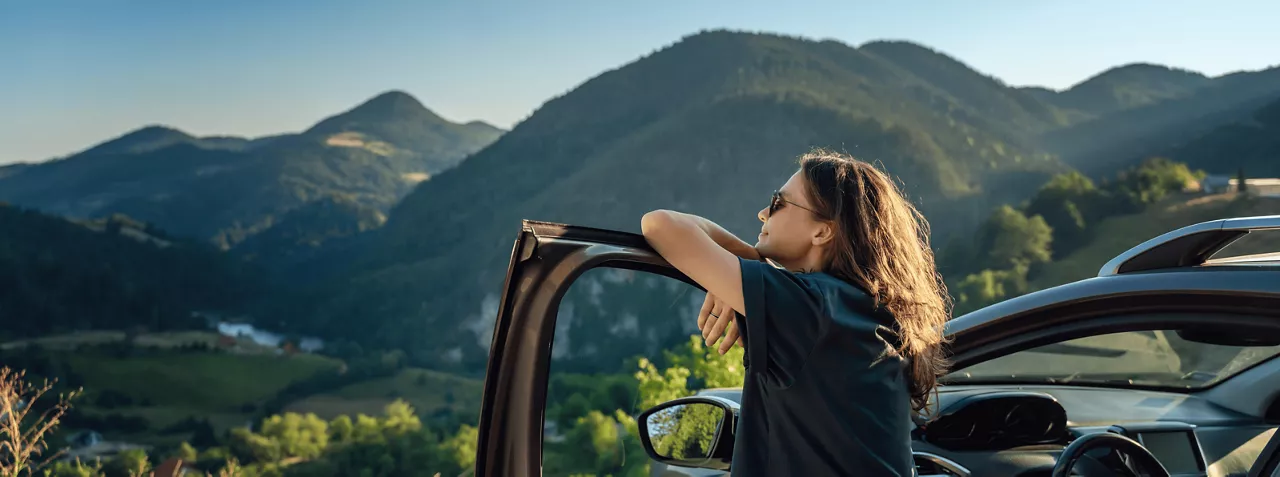  Kobieta w okularach przeciwsłonecznych oparta o otwarte drzwi samochodu z Grupy Volkswagen, patrząca na górski krajobraz.